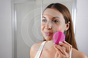 Close up of young woman cleaning skin with silicone brush ultrasonic vibrator in the bathroom. Skin care and wellness concept