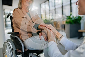 Close-up of young woman doctor holding hands of patient on wheelchair, consoling her.