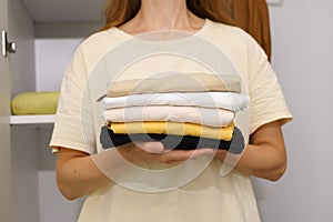 Close-up of a young woman holding a compactly folded stack of clothes for storage against the background of a white closet. The