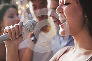 Close- up of young woman holding a microphone and singing at karaoke, friends singing in the background