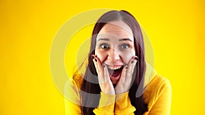 Close up of young woman leaning hands against her cheeks in surprise on yellow background. Pretty female in yellow