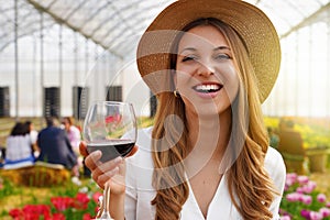 Close-up of young woman looking at camera and holding glass of wine with people among flowers field drinking and chatting on the