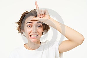 Close up of young woman making fun of friend, smiling and showing loser gesture, l letter on forehead, standing over