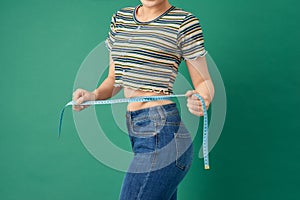 Close up young woman measuring her waist with tape measure over green background