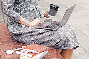 Close up of young woman`s hand holding a cup of coffee while using a laptop on the bench.