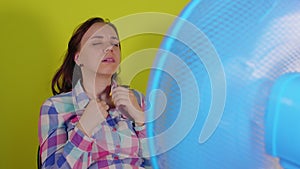 Close up of young woman sitting on chair in front of fan on yellow background. Exhausted brunette escapes from heat