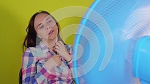 Close up of young woman sitting on chair in front of fan on yellow background. Exhausted brunette escapes from heat