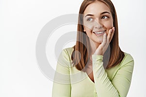 Close up of young woman smiling, touching clean glowing skin and gazing away at promotion text, daydreaming, concept of