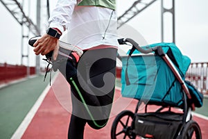 Close up of young woman in sportswear warming up her legs, standing on the bridge with a stroller on a cloudy day in the