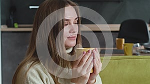 Close-up young woman in sweater enjoys delicious hot tea while sitting on cozy sofa at home. Portrait of beautiful