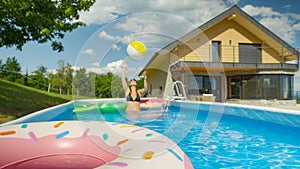 CLOSE UP: Young woman throws an inflatable ball at other floaties in pool.