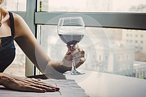 Close up of young woman which her hand holding with glass red wine as a looking out on the window. large glass of red