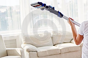 Close up of young woman in white shirt holding in hands wireless vacuum cleaner while cleaning in living room at home, back view.