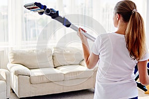 Close up of young woman in white shirt holding in hands wireless vacuum cleaner while cleaning in living room at home, back view.
