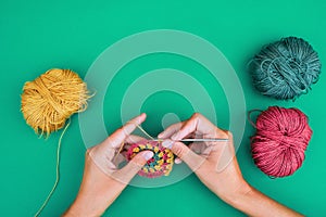 Close view of female hands, making a pattern with crochet hook on green background with multicolored balls of yarn near.