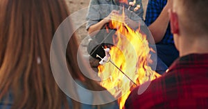 Close view of friends roasting marshmallows sitting around bonfire on sandy beach