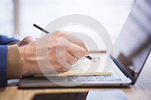 In close view, a man's hand writing down notes in a notepad on a laptop