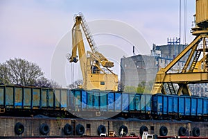 Close view of river port crane loading open-top gondola cars of freight train on cloudy day. Empty river drag boats or