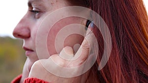 Close of the young girl in a warm red sweater inserts a wireless earphone into her ear. Listening to your favorite music