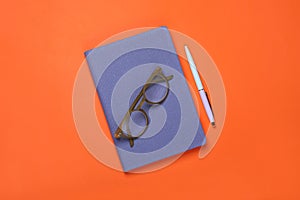 Closed blue notebook, eyeglasses and pen on orange background, flat lay