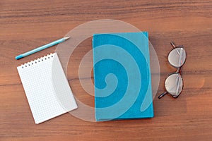 Closed book, eyeglasses, notepad and pen on wooden background