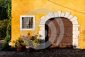 Closed brown wooden entrance door with barred window on yellow wall of ancient medieval village. Strassoldo Italy.