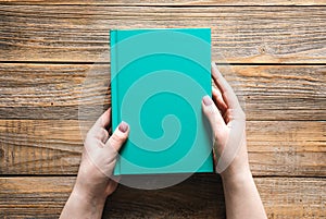 Closed green book in female hands on a wooden background, top view.