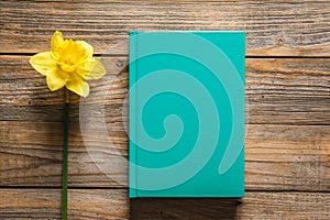 Closed green book and yellow daffodil flower on wooden background, top view.