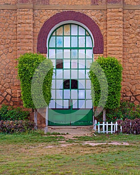 Closed grunge door with green metal grid framed by two green bushes in orange colored bricks stone wall in sunrise time