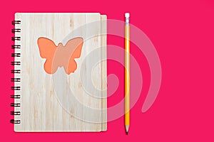 Closed notebook in a bamboo cover with an orange butterfly on a pink background and a pencil