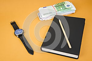 closed notepad, clock, cash and pencil laying on it on office orange table