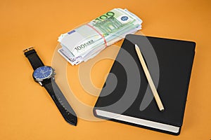 closed notepad, clock, cash and pencil laying on it on office orange table