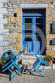 Closed sidewalk cafe with chairs tilted onto small bistro table in front of brightly painted doors in old rock building