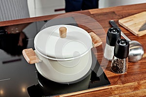 Closed white saucepan with wood pens on the black stove and glass spice mills with papper and salt on the wood table in the