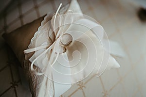 Two gold wedding rings resting on top of a white satin pillow, tied with a white ribbon bow