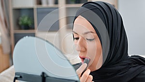 Closeup Arabic woman in black hijab applying lipstick in front of mirror use moisturizer lip gloss