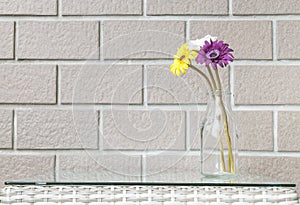 Closeup artificial colorful flower on transparent glass bottle on wood weave table on blurred brown brick wall texture background