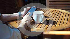 Closeup of Asian male hands playing smartphone and drinking coffee at an outdoor table with morning sunlight.