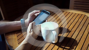 Closeup of Asian male hands playing smartphone and drinking coffee at an outdoor table with morning sunlight