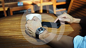 Closeup of Asian male hands playing smartphone and drinking coffee at an outdoor table with morning sunlight.