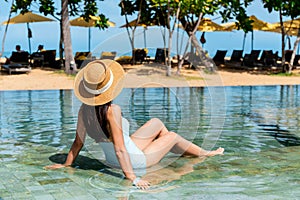 Closeup of Asian woman wear a hat and swimsuit sitting and relaxing in swimming pool at luxury hotel, beach scenery on background
