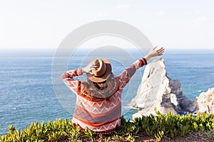 Closeup back view of woman in travel clothes and hat sitting and looking at blue ocean and sky