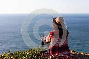 Closeup back view of woman in travel clothes and hat sitting and looking at blue ocean and sky
