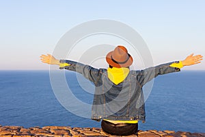 Closeup back view of woman in travel clothes and hat sitting and looking at blue ocean and sky