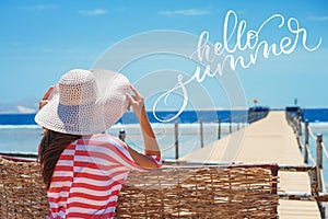 Closeup back view of woman in white hat looking out towards blue ocean and sky and words Hello summer. Calligraphy