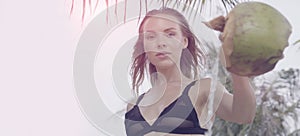 Closeup of beautiful glamour woman wearing black swimwear pouring water from coconut on a beautiful summer day between palm trees