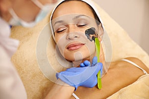 Closeup of beautiful woman`s face and aesthetician`s hands applying a black beauty product on her face