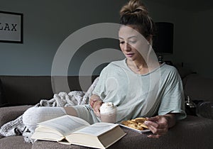 Closeup of beautiful young mixed race, Asian Caucasian, woman reading book at home on couch with hot chocolate milk and cookies