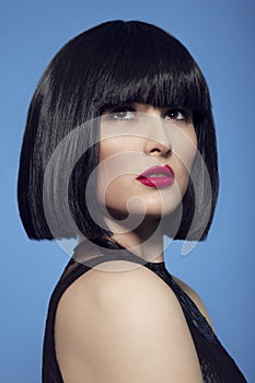 Closeup beauty portrait of a brunette woman in black wig with make up, red lips, over blue background.