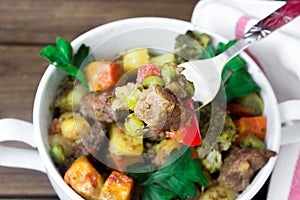Closeup beef meat stewed with vegetables in ceramic pot with a piece on a fork on wooden background